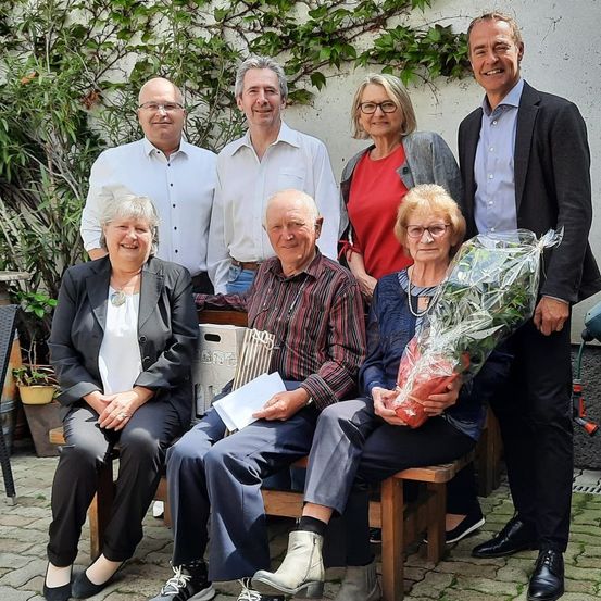 Eine Gruppe von sieben Personen sitzt auf einer Bank und lächelt für ein Foto. Zwei Frauen halten Blumensträuße, ein Mann liest ein Papier. Hinter ihnen stehen zwei Männer mit einer Frau in der Nähe einer Wand mit Kletterpflanzen.