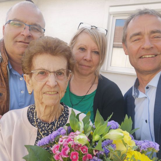 Ein Gruppenfoto mit einer älteren Frau, die einen Blumenstrauß hält. Hinter ihr stehen drei Personen: ein Mann mit Brille, eine Frau mit einer Brille auf dem Kopf und ein weiterer Mann. Sie stehen vor einer weißen Wand mit einem Fenster.