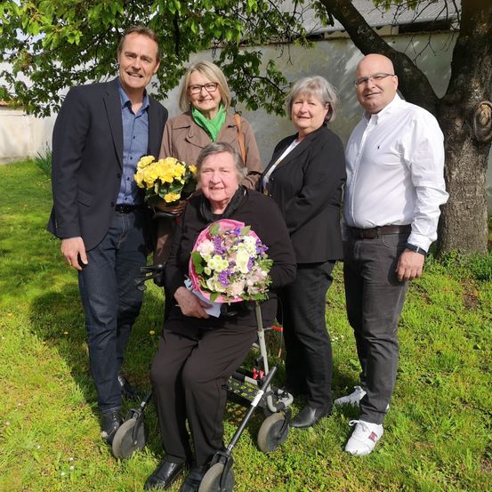 Fünf Menschen posieren für ein Foto auf der Rasenfläche. Eine Frau sitzt auf einem Rollator und hält einen Blumenstrauß. Eine andere Frau, die neben ihr steht, hält einen weiteren Blumenstrauß. Ein Mann steht neben ihnen. Dahinter ist ein Baum gepflanzt.