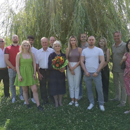 Eine Gruppe von Menschen steht im Freien unter einem Weidenbaum und lächelt für ein Foto. Eine Frau hält einen Blumenstrauß. Sie tragen Freizeitkleidung und der Hintergrund ist unscharf.