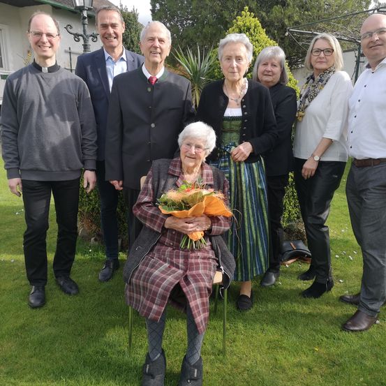 Eine Gruppe von Menschen steht in einem Garten. Eine ältere Frau sitzt in der Mitte und hält einen Blumenstrauß. Sie sind feierlich gekleidet, und einige tragen Brillen. Im Hintergrund befinden sich Grünpflanzen und ein Gebäude.