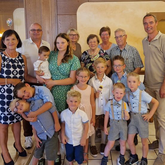 Eine Familie von 14 Personen posiert für ein Foto in einem Raum. Sie alle lächeln und scheinen einen besonderen Anlass zu feiern. Sie tragen Freizeitkleidung und Turnschuhe. Einige von ihnen halten Eis und tragen Brillen. Der Raum hat Holzwände und geflieste Böden.