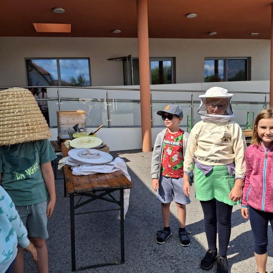 Vier Kinder stehen draußen, zwei in Imkeranzügen. Eines trägt einen Strohhut. Ein Holztisch hat Teller und Tücher. Ein Gebäude mit Glasfenstern und einem Metallzaun steht dahinter.