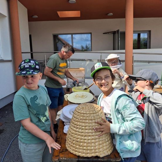 Eine Gruppe von Kindern und ein erwachsener Mann sind vor einem Gebäude. Der Mann trägt einen Imkeranzug und kümmert sich um einen Bienenstock. Die Kinder beobachten.