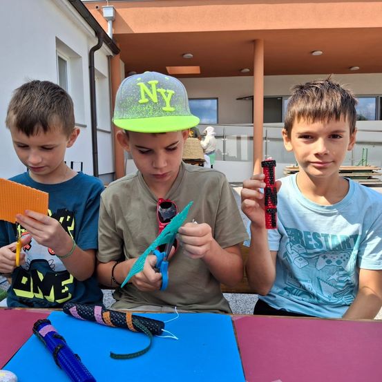 Drei Jungen sitzen um einen Tisch herum und beschäftigen sich mit Bastelarbeiten. Ein Junge hält einen kleinen, bunten Gegenstand, ein anderer ein gelbes Objekt und der dritte ein farbiges Papierstück. Sie sind von verschiedenen Bastelmaterialien umgeben.