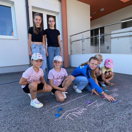 Eine Gruppe junger Mädchen ist auf dem Boden, einige knien und zeichnen auf den Beton. Sie tragen Freizeitkleidung und einige haben Mützen. Hinter ihnen steht ein Gebäude mit Glasfenstern und einer Balustrade.
