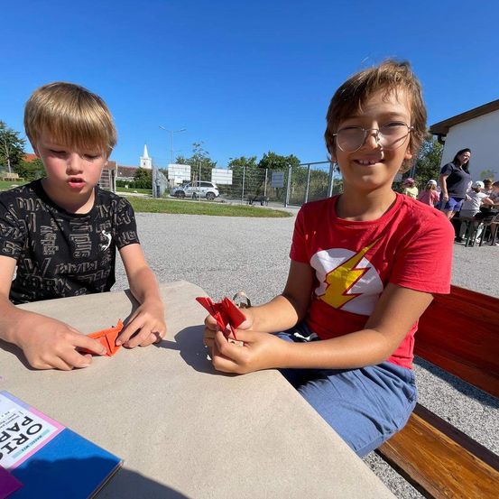 Zwei Jungen sitzen an einem Tisch, lächeln und halten jeweils ein orangefarbenes Papierflugzeug in den Händen. Sie scheinen einen sonnigen Tag draußen zu genießen, möglicherweise in einem Park.