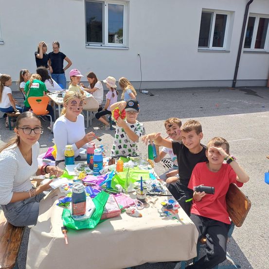 Eine Gruppe von Kindern und Erwachsenen sitzt um einen Tisch herum, auf dem verschiedene Kunstmaterialien wie Farben und Pinsel stehen. Einige von ihnen bemalen ihre Gesichter. Die Umgebung ist im Freien vor einem Gebäude.