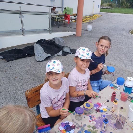 Drei Mädchen sitzen draußen an einem Tisch und beschäftigen sich mit einer Malaktivität. Sie haben Malpinsel und Flaschen mit Farbe vor sich. Dahinter steht ein Gebäude mit einer Bank und einem Tisch mit einigen Gegenständen darauf.
