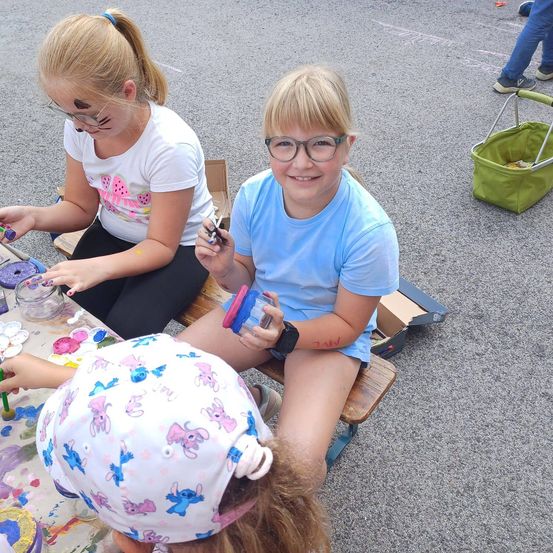 Drei Mädchen sitzen auf einer Bank und beschäftigen sich mit einer Malaktivität. Eine hält einen Pinsel, eine andere einen Glaskrug und die dritte hat ein bemaltes Gesicht. Dahinter steht eine Person mit einem grünen Korb.