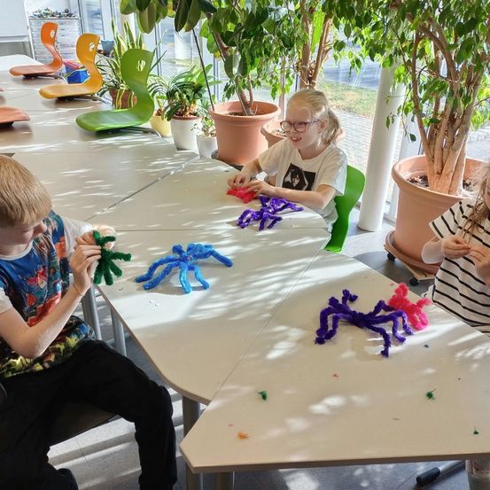 Drei Kinder arbeiten am Tisch an der Herstellung von Pfeifenreiniger-Spinnen, wobei ein Junge in einem bunten Shirt und ein Mädchen in einem weißen Shirt ist. Der Raum hat große Topfpflanzen und helles Sonnenlicht.