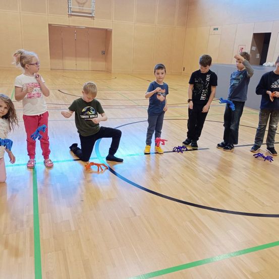 Eine Gruppe von Kindern spielt ein Spiel mit spinnenartigen Spielzeugen auf einem Holzboden einer Turnhalle. Sie stehen in einer Reihe, jeder hält ein Spielzeug.