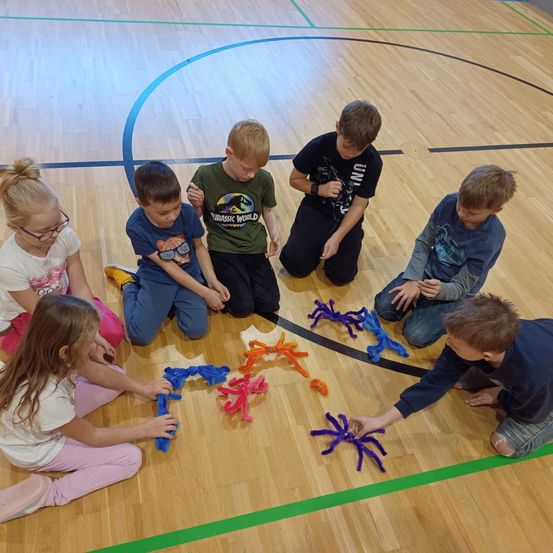Eine Gruppe von Kindern spielt mit farbenfrohen Pipe-Cleaner-Spinnen auf einem Basketballplatz. Sie sitzen im Kreis und halten jeweils eine Spinne.