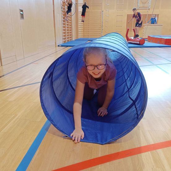 Ein junges Mädchen kriecht durch einen blauen Tunnel in einer Turnhalle mit anderen Kindern. Sie trägt eine Brille und ein braunes Shirt. Hinter ihr beschäftigen sich andere Kinder mit verschiedenen Aktivitäten.