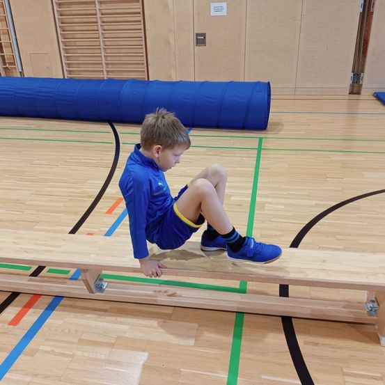 Ein Junge sitzt auf einer Holzbank in einer Turnhalle und macht möglicherweise eine Übung. Er trägt ein blaues Outfit.