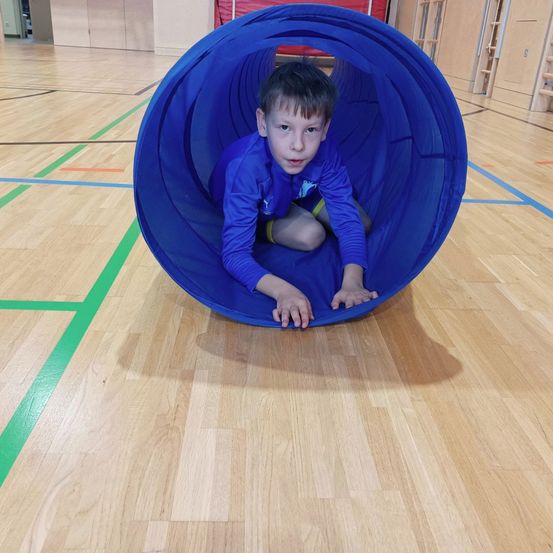 Ein junger Junge kriecht durch einen großen blauen Tunnel auf einem Turnhallenboden, umgeben von Holzpaneelen und bunten Markierungen.