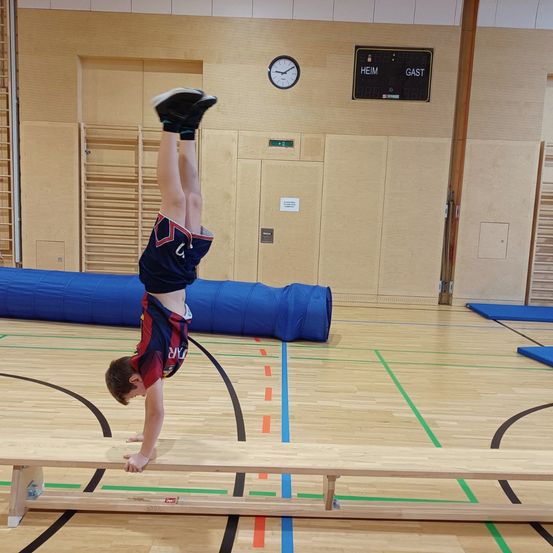 Ein junger Junge balanciert auf dem Kopf auf einer hölzernen Bank in einer Turnhalle. Er trägt ein blaues und rotes Sporttrikot. Hinter ihm liegt ein blauer aufblasbarer Schlauch auf dem Boden.