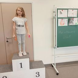 Ein kleines Mädchen mit blonden Haaren steht auf einem Podest mit den Nummern '1' und '3'. Sie trägt ein graues T-Shirt mit 'CHEF' darauf, graue Hosen und pinke Schuhe. Hinter ihr befindet sich eine Tür, und eine Tafel mit Zeichnungen und dem Text 'Malwettbewe 2025' steht rechts.