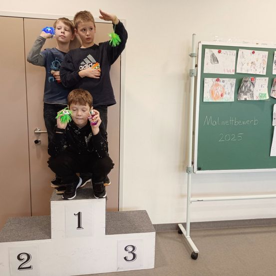 Drei Jungen stehen auf einem Podest, wobei der Junge in der Mitte Spielzeug hält. Sie sind drinnen, in der Nähe einer Tür und einer Tafel mit dem Text 'Malwettbewerb 2025.'