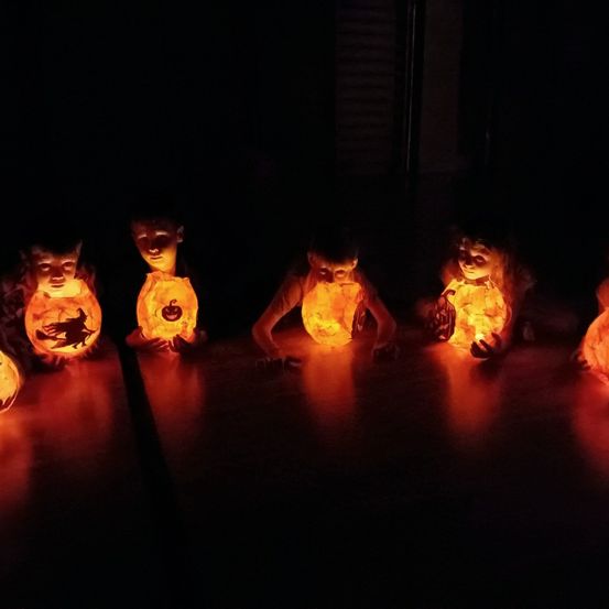 Fünf Kinder sitzen in einer Reihe und halten leuchtende Laternen, ihre Gesichter vom warmen Licht beleuchtet. Die Umgebung ist dunkel, nur ihre Laternen spenden Licht.