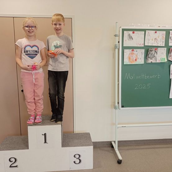 Zwei Kinder stehen lächelnd auf einem Podest. Das Mädchen trägt ein weißes T-Shirt mit Herzdesign und eine Brille, während der Junge ein graues T-Shirt trägt. Sie stehen vor einer grünen Tafel mit Zeichnungen und der Nummer 2025.