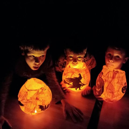 Drei Kinder, beleuchtet von Kürbislaternen, schauen angestrengt in die Dunkelheit. Jede Laterne hat ein unterschiedliches Halloween-Design.