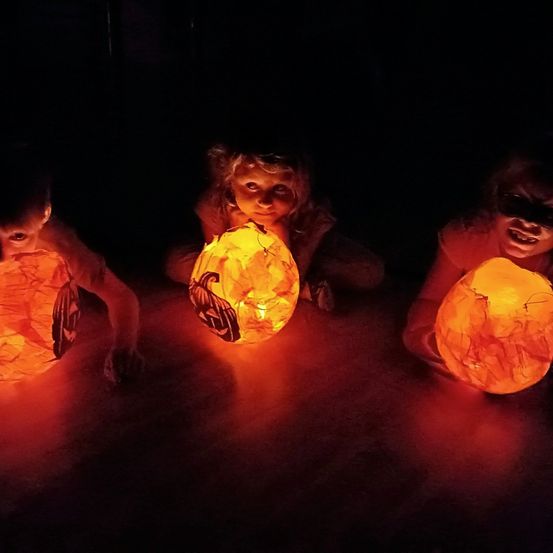 Drei Kinder sind in einem dunklen Raum und halten leuchtende Kürbisse. Sie lächeln fröhlich. Der Raum ist dunkel und nur die Kürbisse spenden Licht.