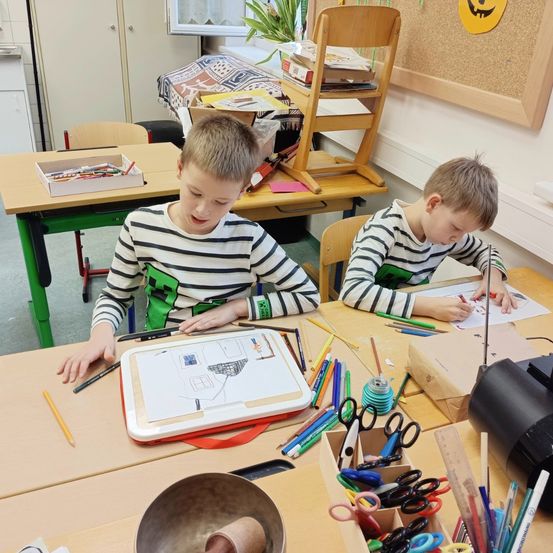 Zwei junge Jungen sitzen an einem Schreibtisch im Klassenzimmer. Sie zeichnen mit farbigen Stiften und haben eine Schachtel mit Scheren in der Nähe.