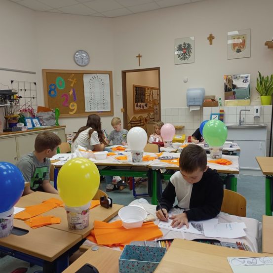 Ein Klassenzimmer mit Kindern, die an einem Bastelprojekt teilnehmen. Die Tische sind mit Luftballons, Papieren und verschiedenen Bastelutensilien dekoriert. Ein Bulletin-Board mit Zahlen und Bildern im Hintergrund.
