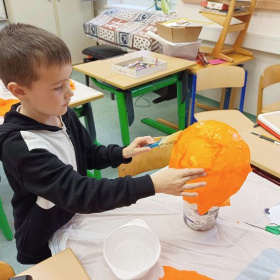 Ein junger Junge malt einen orangefarbenen Ballon in einem Klassenzimmer, umgeben von Tischen und Stühlen.
