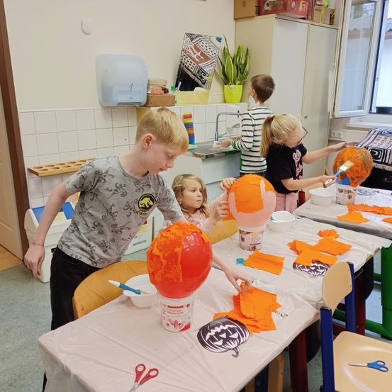 Vier Kinder malen Kürbisse in einem Raum mit Tischen und Stühlen. Sie verwenden orangefarbene Farbe und es gibt Schalen mit Farbe und Pinsel auf den Tischen.