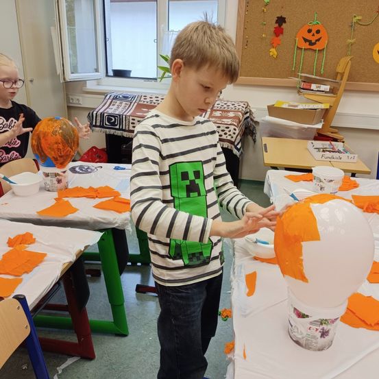 Ein Junge malt eine Kürbis in einem Klassenzimmer mit Halloween-Dekorationen. Ein anderes Kind malt einen Kürbis im Hintergrund.