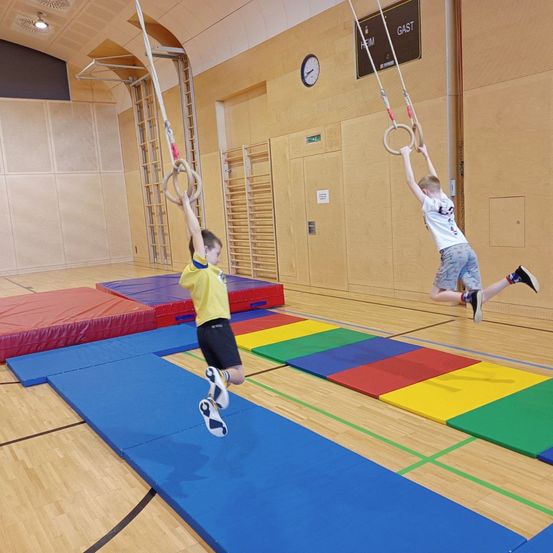 Zwei Kinder schwingen an Ringen in einer Turnhalle, eines springt von einer blauen Matte auf eine andere. Bunte Matten bedecken den Boden, und eine Wand mit einer Uhr und einem Schild befindet sich dahinter.