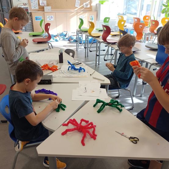 Vier Jungen sitzen an einem Tisch in einem Klassenzimmer und beschäftigen sich mit dem Basteln von Spinnen mit Rohrreinigern. Ein Junge konzentriert sich auf den Zusammenbau seiner Spinne, während ein anderer ein fertiges Werk in den Händen hält. Das Klassenzimmer ist mit Stühlen und Tischen gefüllt.