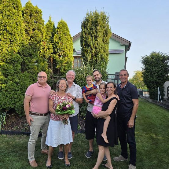Eine Familie mit sieben Personen steht in einem Garten vor einem Haus. Sie lächeln und scheinen für ein Foto zu posieren. Dahinter befinden sich Sträucher und ein Zaun.