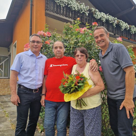 Vier Personen stehen vor einem Haus mit Blumen. Der Mann links trägt ein blaues Hemd, während die zentrale Figur, eine Frau, einen Blumenstrauß hält. Der Mann rechts trägt ein graues Poloshirt.
