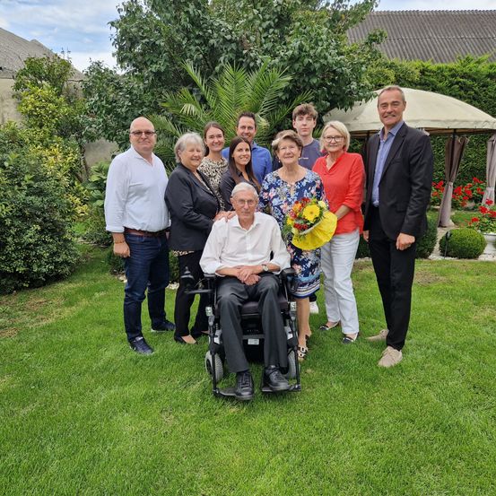 Eine Gruppe von Menschen steht in einem Garten. Ein Mann im Rollstuhl hält Blumen. Im Hintergrund befindet sich ein Zelt und ein Haus.