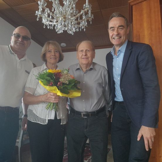 Vier Personen stehen lächelnd zusammen. Eine Frau hält einen Blumenstrauß. Der Innenraum ist mit einer Kronleuchter und einer Holzwand zu sehen.