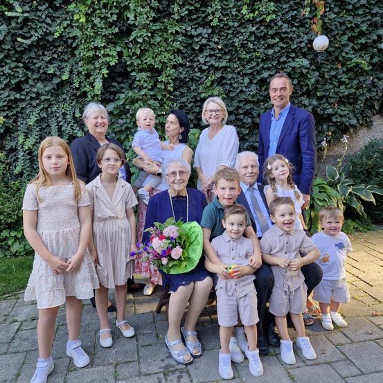 Eine große Familie posiert für ein Foto im Garten, mit einer grünen Wand aus Efeu im Hintergrund. Die Familie besteht aus Kindern, Erwachsenen und einem Baby, das gehalten wird.
