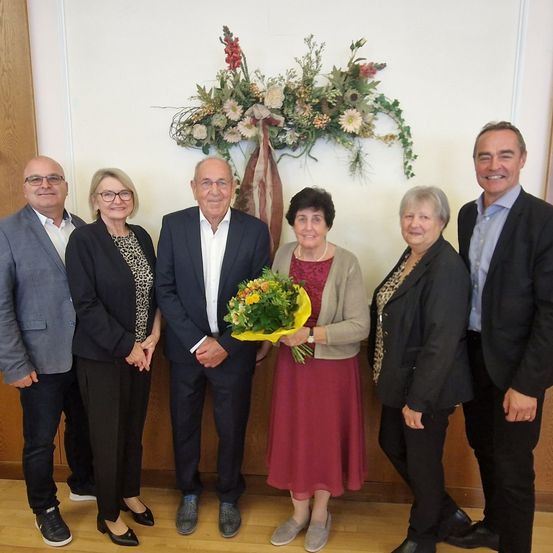 Eine Gruppe von sechs Personen steht in einem Raum und lächelt für ein Foto. Eine Frau hält einen Blumenstrauß. Hinter ihnen schmückt ein Blumenarrangement die Wand.