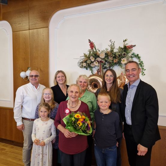 Eine Gruppe von Menschen posiert für ein Foto vor einer weißen Wand mit einem Blumenarrangement. Eine ältere Frau hält einen Blumenstrauß. Es sind Kinder und Erwachsene in formeller Kleidung anwesend.