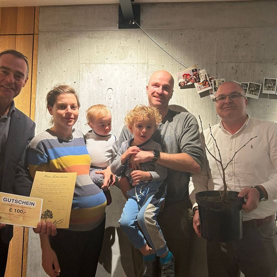 Eine Familie mit zwei kleinen Kindern und zwei Männern hält ein Zertifikat und einen Topf mit Pflanzen und lächelt für ein Foto vor einer Wand mit Fotos.
