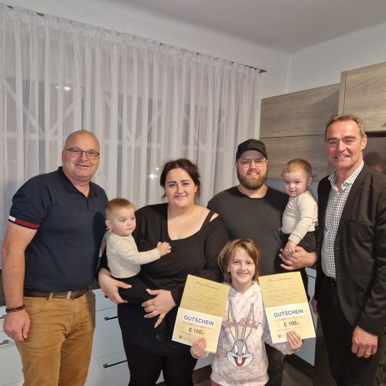 Eine Familie mit fünf Personen, darunter zwei kleine Kinder, steht in einer Küche. Sie halten Gutscheine mit dem Text 'Gutschein' und 'C 100-' in den Händen. Die Kinder lächeln.