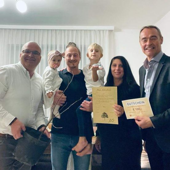 Fünf Erwachsene und zwei Kleinkinder posieren drinnen mit Zertifikaten. Der Mann links hält eine Topfpflanze. Der Mann in der Mitte hält zwei Kleinkinder, eines mit einem Schnuller. Die Frau rechts hält ein Zertifikat mit einem Geschenkgutschein.