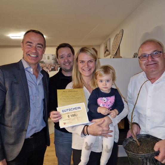 Eine vierköpfige Familie posiert drinnen für ein Foto. Ein Mann hält einen Geschenkgutschein, eine Frau eine kleine Mädchen und ein weiterer Mann einen Topf mit Pflanze. Sie alle lächeln für die Kamera.