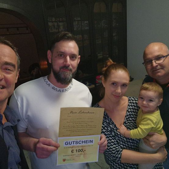 Ein Familienfoto mit einem Mann, der ein Zertifikat für einen Geschenkgutschein im Wert von 100 Euro hält. Der Mann und die Frau lächeln, während das Baby neugierig schaut. Sie sind in einem Raum mit einer warmen Atmosphäre.