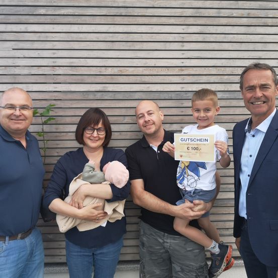 Fünf Erwachsene und Kinder stehen lächelnd vor einer Holzwand. Eine Frau hält ein Baby, ein Junge ein Zertifikat.