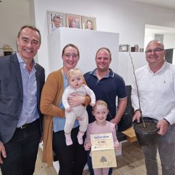 Fünf Erwachsene und zwei Kinder posieren für ein Foto. Eine Frau hält ein Baby und ein Zertifikat, und ein Mann hält eine Topfpflanze.