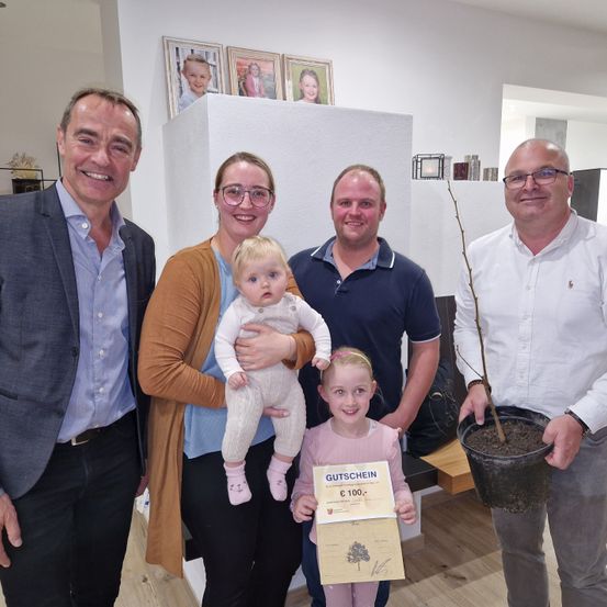 Fünf Erwachsene und zwei Kinder posieren für ein Foto. Eine Frau hält ein Baby und ein Zertifikat, und ein Mann hält eine Topfpflanze.