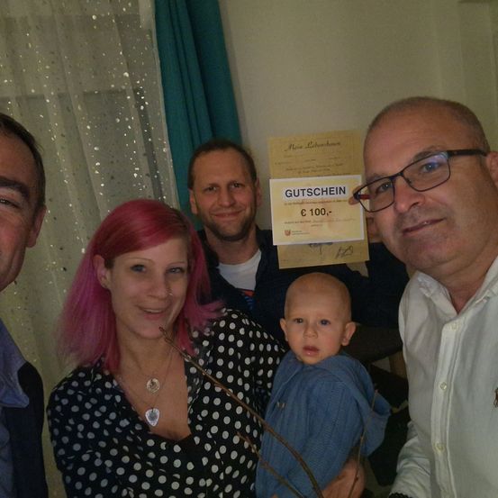 Eine Gruppe von Erwachsenen und einem Baby lächelt in einem Raum. Ein Mann hält ein Zertifikat und einen Stock.